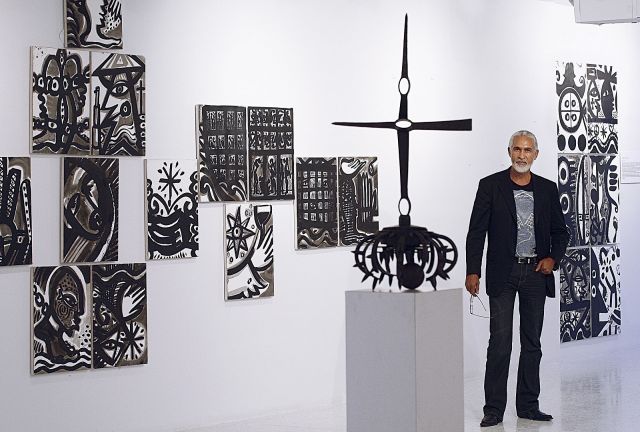 A middle-aged Haitian man in a dark suit jacket and jeans stands in front of an exhibit of his black and white abstract artwork