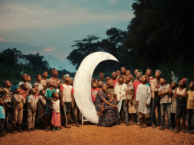 A woman sits in a carved wooden crescent moon.  She's holding her child, and another 30 children are gathered around her