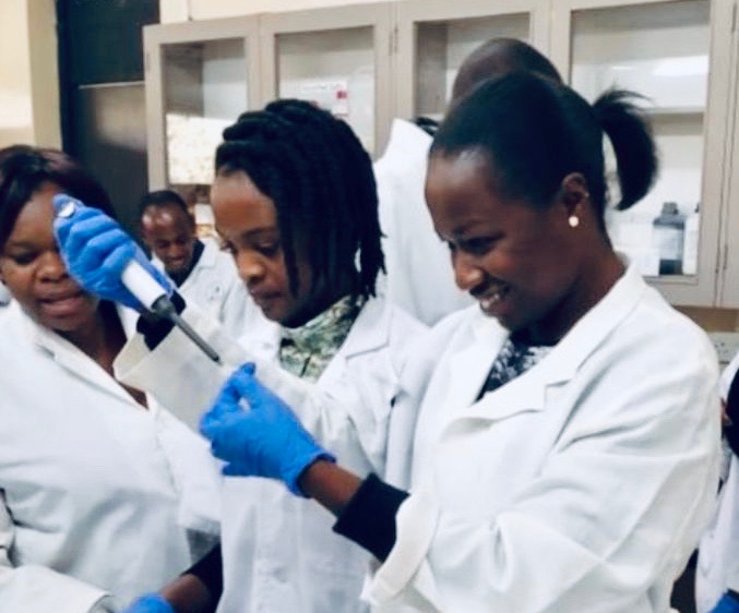 A Kenyan woman in a white lab coat and blue gloves scrapes a sample off a microscope slide